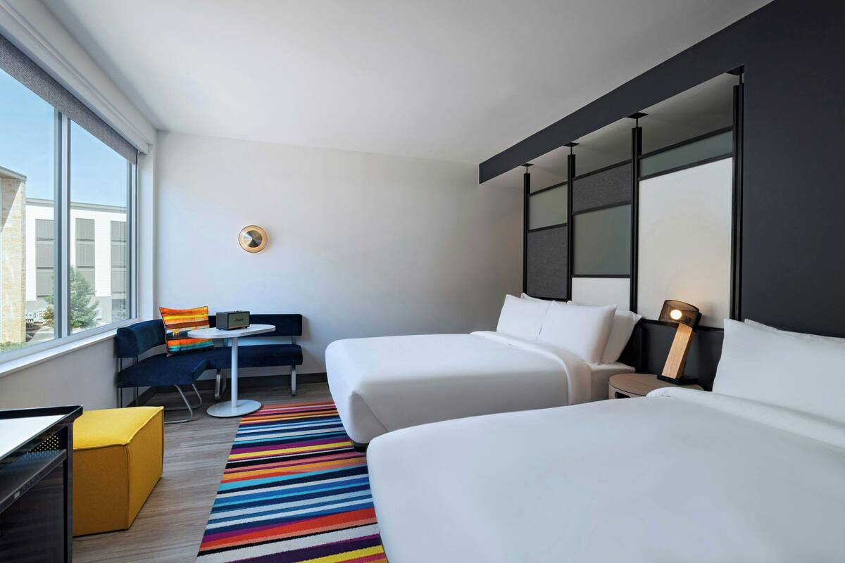 A hotel room with two beds, a desk, a chair, a small table, and a colorful striped rug.