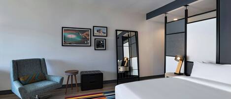 A modern hotel room with a bed, a chair, a small table, a colorful rug, and wall-mounted artwork.