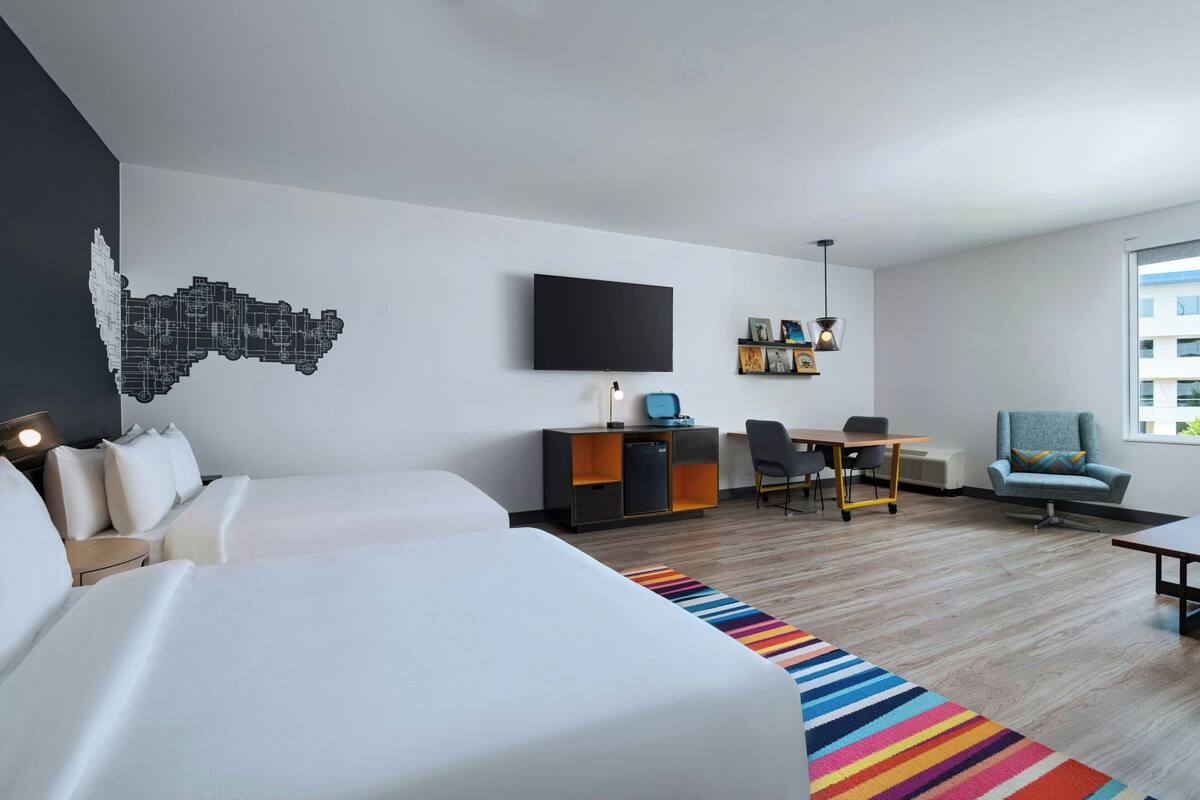 A hotel room with two beds, a desk, a chair, a TV, and a window.