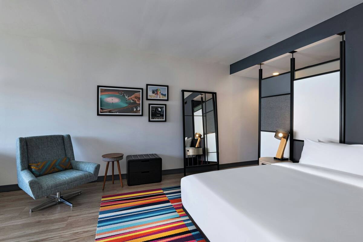A modern hotel room with a large bed, a grey armchair, a colorful striped rug, and a mirror with a black frame.