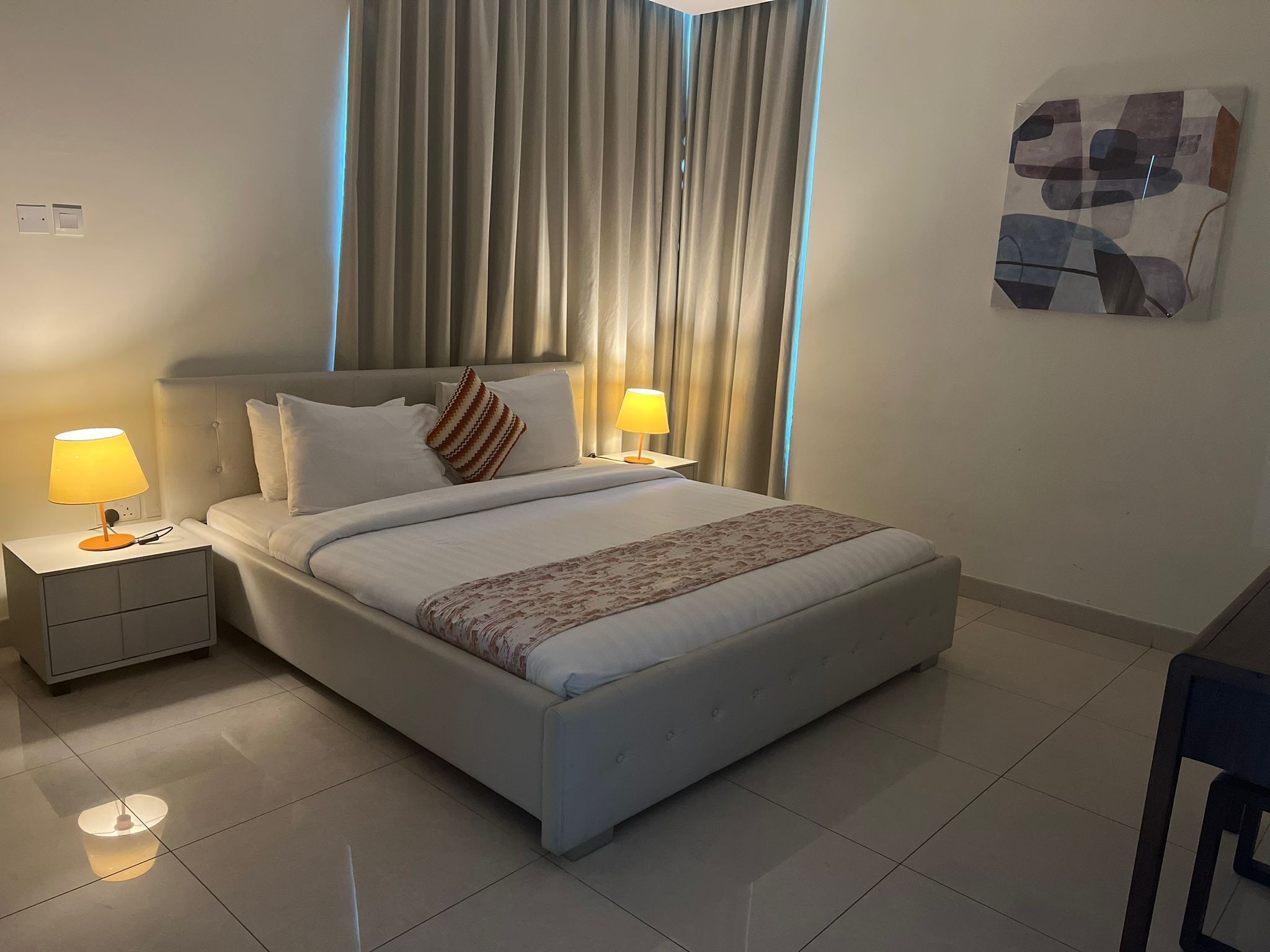 A neatly made bed with a patterned throw, two bedside lamps, a headboard, and a large window with curtains.
