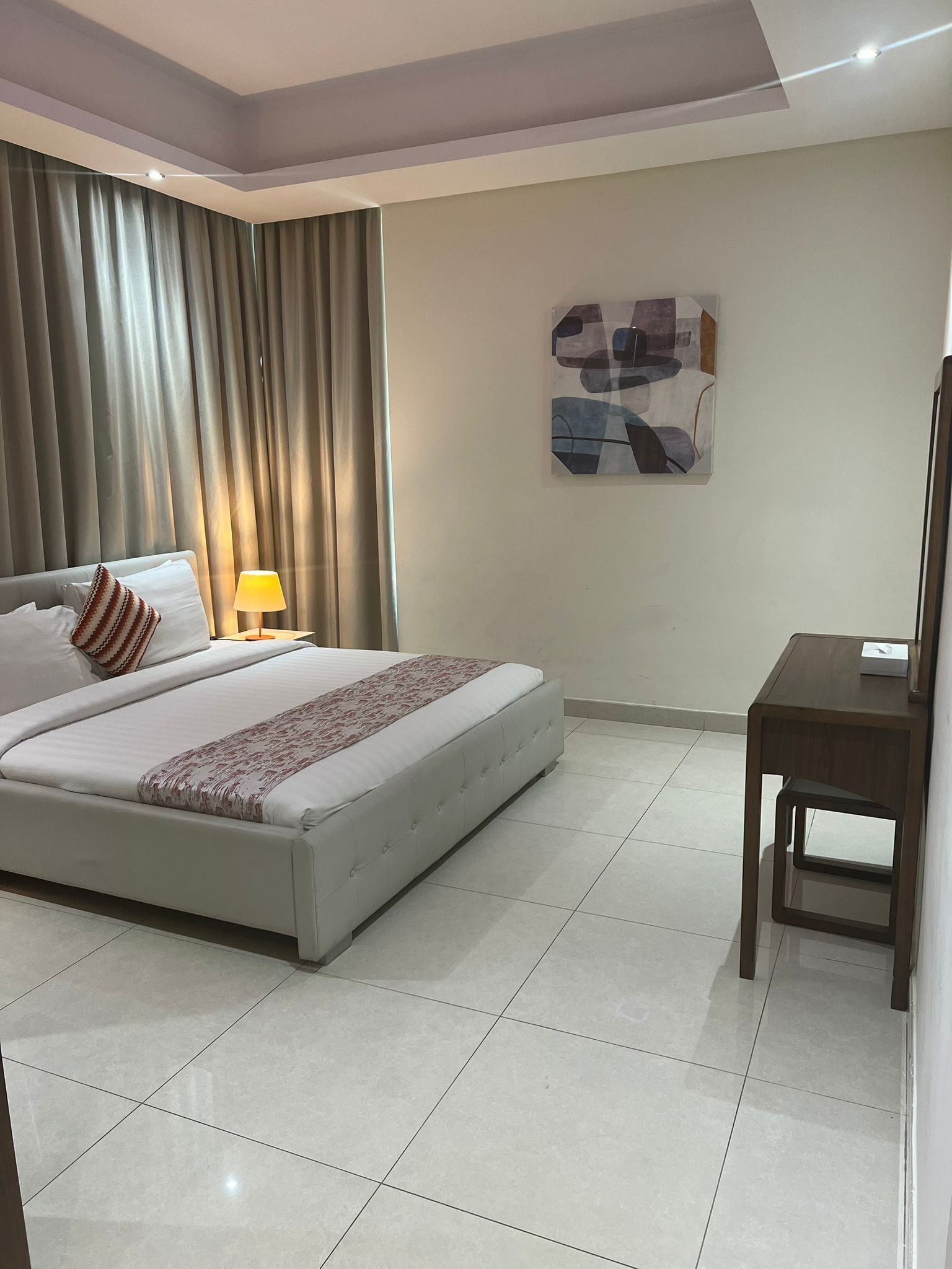 A hotel room with a bed, a bedside lamp, a small table, and a painting on the wall.
