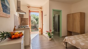 Ein gemütliches Schlafzimmer mit Bett, Holzschrank, einer Küchenzeile mit Spüle und einem Balkon mit Blick auf Bäume.