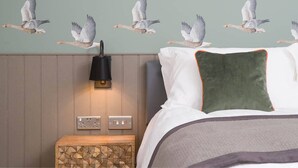 A bedroom with a bed, pillows, a nightstand, a wall-mounted lamp, and a wall with a bird mural. - The Chequers (Knebworth)