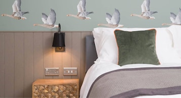 Quarto com cama, almofadas, mesa de cabeceira, luminária na parede e um mural de pássaros na parede.
