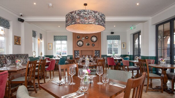 A spacious dining area with multiple wooden tables and chairs, a large floral-patterned ceiling light, and framed artwork on the walls. - The Chequers (Knebworth)