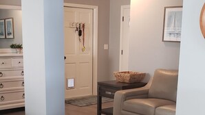Ett vardagsrum med en beige soffa, ett soffbord och ett trägolv. Det finns en dörr som leder till ett annat rum med en vit vägg och en inramad tavla.