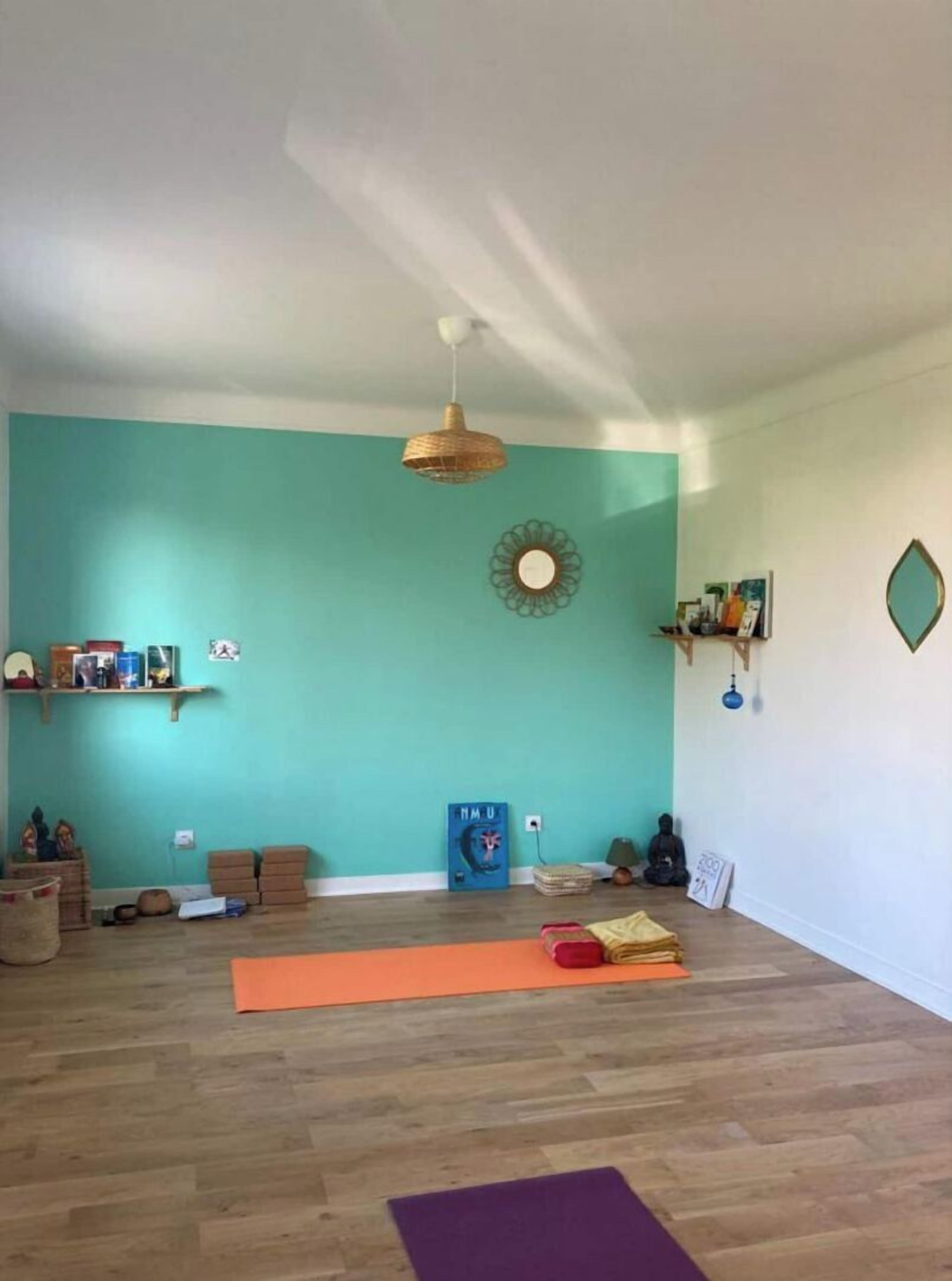 Une pièce spacieuse aux murs de couleur turquoise, dotée d’un plancher en bois et d’un ventilateur de plafond. On y trouve des tapis de yoga, un coussin de méditation et une petite étagère avec des livres et des objets décoratifs.