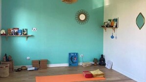 Ein geräumiges Zimmer mit Türkiswänden, Holzböden und einem Deckenventilator. Es befinden sich Yogamatten, ein Meditationskissen und eine kleine Ablage mit Büchern und Dekorationsobjekten dort.