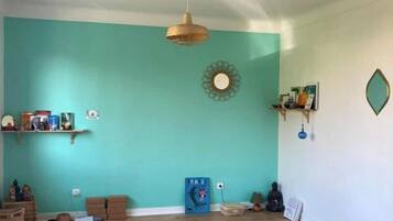 Une pièce spacieuse aux murs de couleur turquoise, dotée d’un plancher en bois et d’un ventilateur de plafond. On y trouve des tapis de yoga, un coussin de méditation et une petite étagère avec des livres et des objets décoratifs.