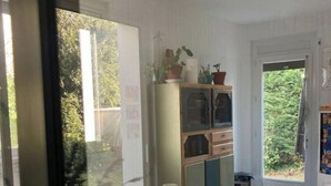 Ein Essbereich mit einem runden Tisch, bedeckt mit einer blumigen Tischdecke, umgeben von Holzsesseln. Ein Schrank mit geöffneten Türen zeigt verschiedene Gegenstände.