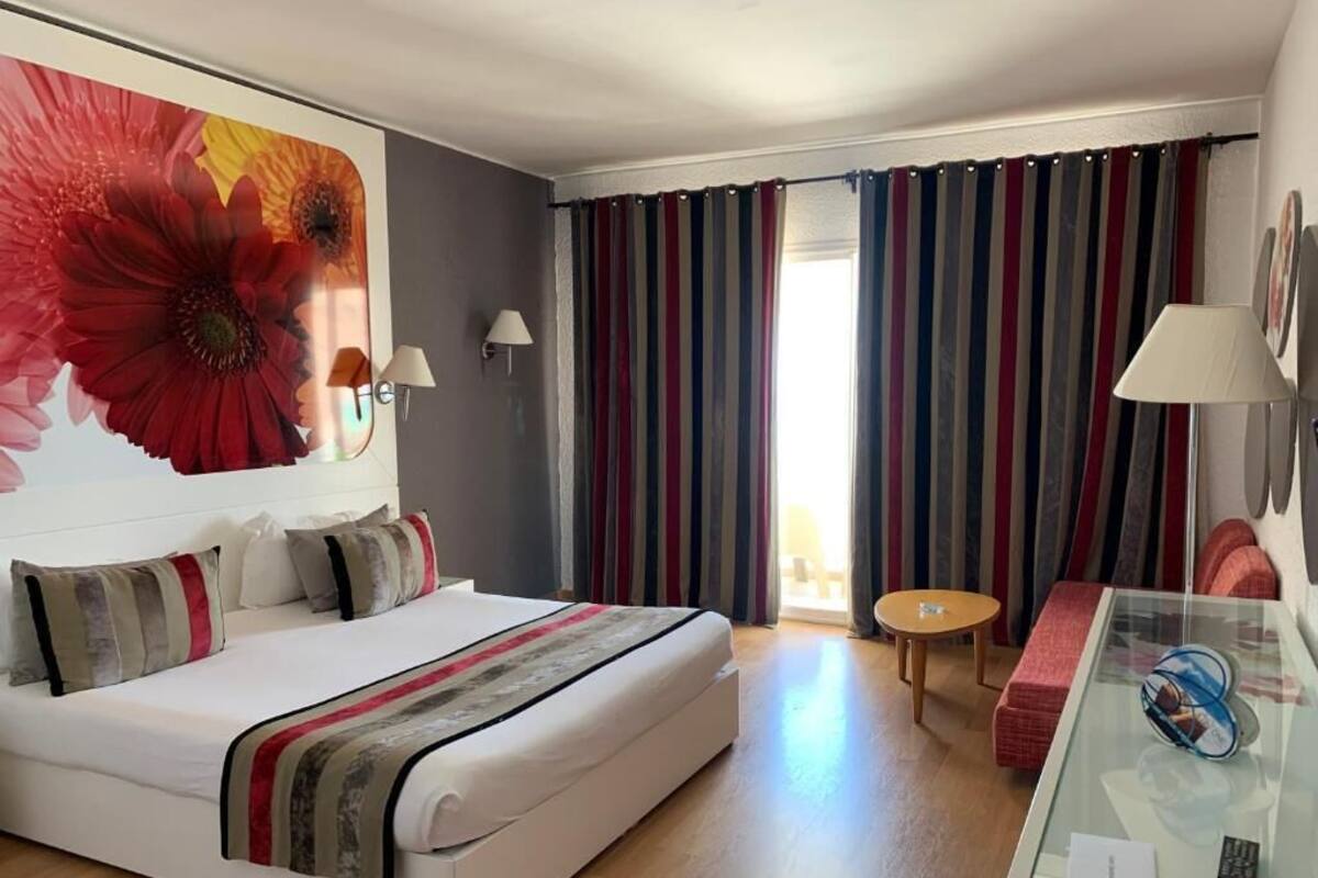 Quarto de hotel com cama, uma mesinha redonda, uma mesa de vidro com um controle remoto e uma janela grande com cortinas.