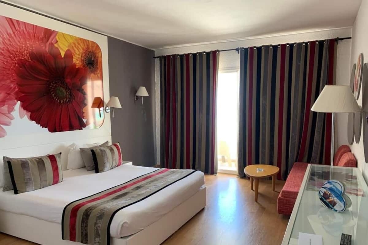 Quarto de hotel com cama, uma mesinha redonda, uma mesa de vidro com um controle remoto e uma janela grande com cortinas.