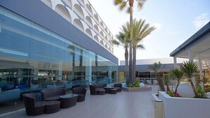 An outdoor seating area with modern furniture, a glass building, and palm trees. - One Resort Jockey (Monastir)