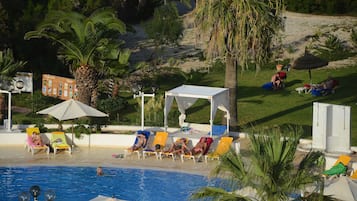 Ein Poolbereich mit Liegestühlen und Sonnenschirmen, umgeben von Palmen und einem Gebäude im Hintergrund.