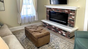Uma sala de estar com lareira, uma TV de tela plana montada acima, um banquinho marrom e um tapete estampado.