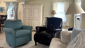 Uma sala de estar com uma poltrona verde-água, uma poltrona de couro preto e um sofá estampado. Há um armário branco com portas decorativas, um abajur e uma janela com cortinas.