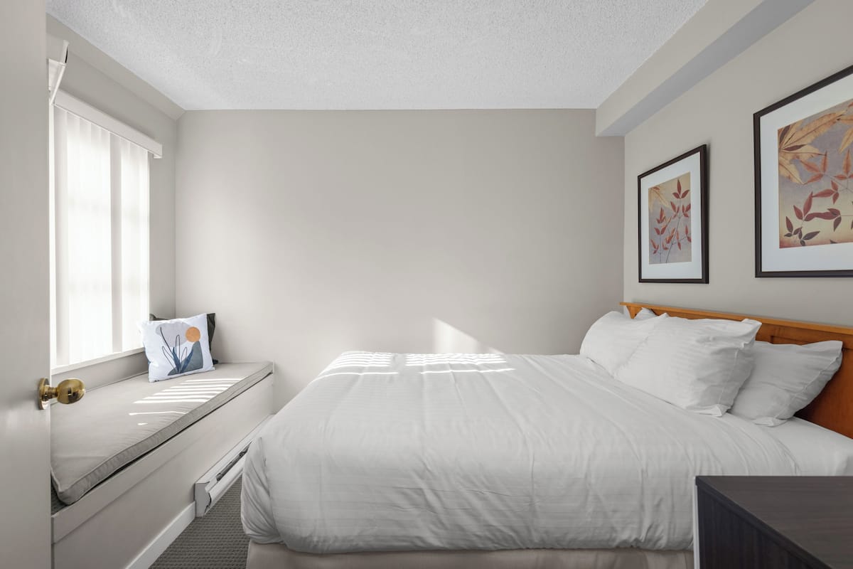 A bedroom with a bed, a window with blinds, a bedside table, and framed artwork on the wall.