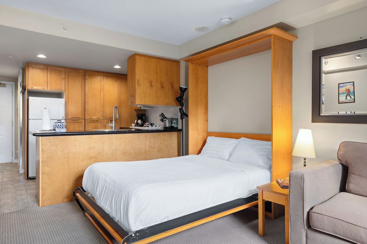 A hotel room with a bed, a small desk, a chair, a kitchenette, and wooden cabinets.