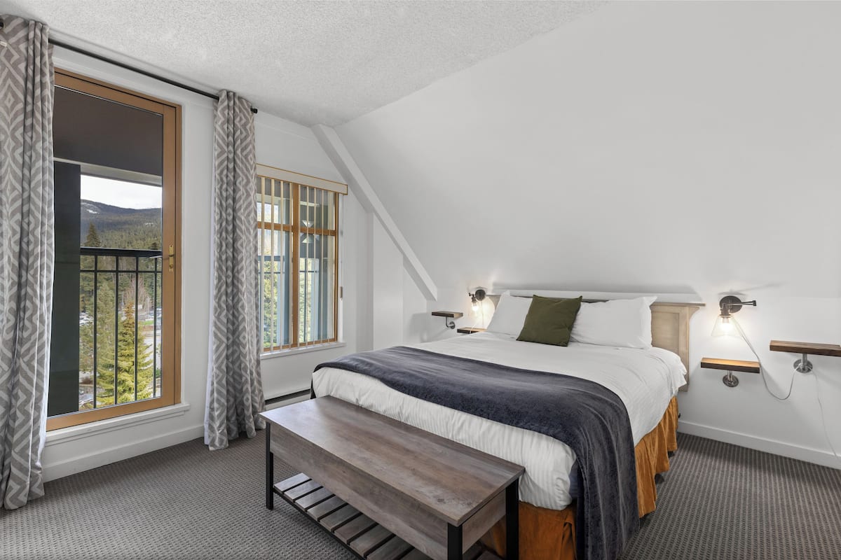 A bedroom with a large bed, a bench, a window with curtains, and a view of trees and mountains.