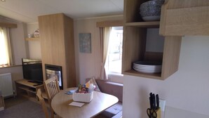 A cozy room with a wooden dining table, a sofa, a TV, and a kitchen area with shelves.