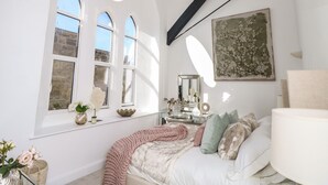 A bedroom with a large arched window, a bed with multiple pillows, a bedside table with a mirror, and a wall-mounted lamp. - Pilgrims Loft (Ventnor)