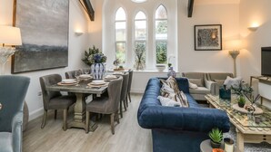 A dining area with a round table set for four, surrounded by upholstered chairs. There are two large windows with arched tops, allowing natural light into the room. A modern sofa and a coffee table with decorative items are visible in the foreground. - Pilgrims Loft (Ventnor)