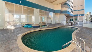 Una piscina interior con paredes de vidrio transparente, un suelo de baldosas y un área junto a la piscina con sillones de descanso.