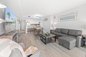 Een moderne woonkamer met een grijze bank, een eethoek met een tafel en stoelen, en een keuken op de achtergrond.