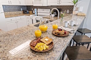 A breakfast setup on a kitchen island with toast, scrambled eggs, bacon, pancakes with berries, and orange juice.