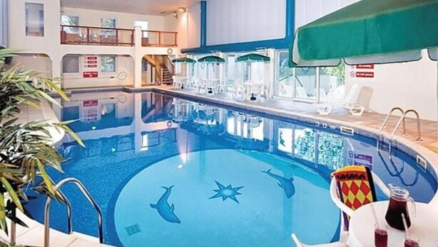 Ein Hallenbad mit Delphinmotiven auf dem Boden, umgeben von Liegestühlen und Sonnenschirmen.