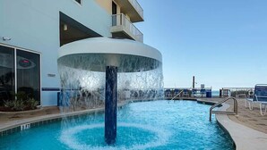 Una piscina con una fuente central, rodeada por una terraza con sillas de descanso y un edificio con balcones.
