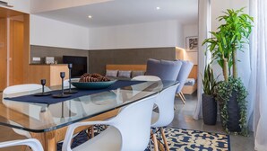 Ein modernes Wohnzimmer mit einem Glastisch, weißen Stühlen, einer blauen Couch mit Kissen sowie einem Couchtisch mit Kiefernzapfen und einer großen Küsterpflanze.