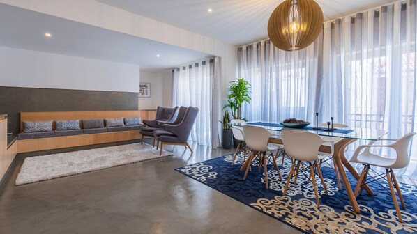 A modern living room with a sofa, a dining table with chairs, a large window with sheer curtains, and a hanging light fixture.