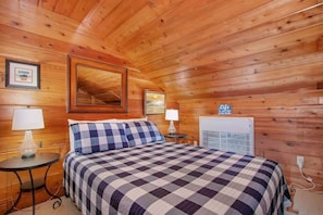 A cozy room with wooden walls and ceiling, a bed with a checkered bedspread, two bedside tables with lamps, and a framed picture on the wall. - See Shore (South Haven)
