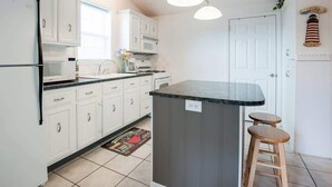 A kitchen with white cabinets, a central island with stools, a microwave, and a clock on the wall. - Sweet Retreat in South Haven • 2 Blocks from Beach (South Haven)