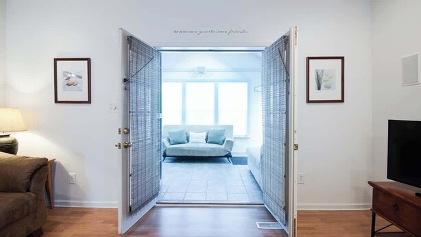 A room with a wooden floor, a sofa, a TV stand, and a door leading to a balcony with a view. - Sweet Retreat in South Haven • 2 Blocks from Beach (South Haven)