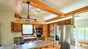 A kitchen with wooden cabinets, a stainless steel refrigerator, a granite countertop, and a ceiling fan. - Driftwood Manor, 6BR, South Haven, Close to Beach (South Haven)
