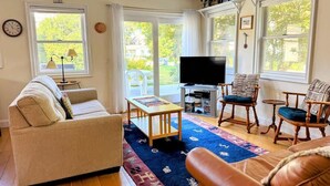 Ein Wohnzimmer mit einer Couch, Sesseln, einem Couchtisch, einem Fernseher und großen Fenstern.