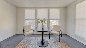 A room with a round table, two chairs, a vase with a plant, and large windows with blinds. - Quaint 1 BR Apt | Mins from ZOO, USC & FT Jackson (Columbia)