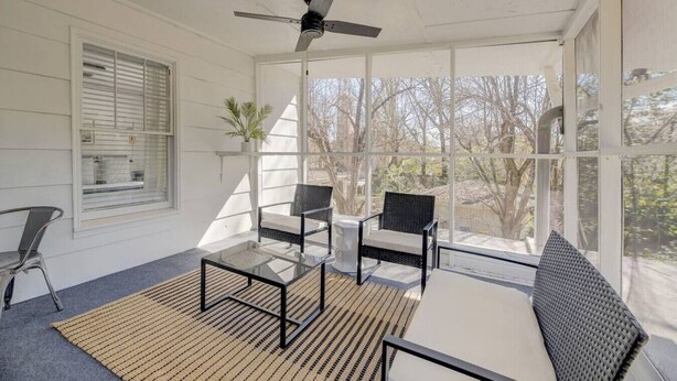 A sunlit room with a ceiling fan, two wicker chairs, a glass table, and a view of trees through large windows. - Quaint 1 BR Apt | Mins from ZOO, USC & FT Jackson (Columbia)