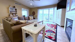A living room with a brown sofa, a small table, and a ceiling fan. There is a window with curtains and a view of the outdoors. - Mariner's Cove 8 • Riverfront • Walk To Beach (South Haven)