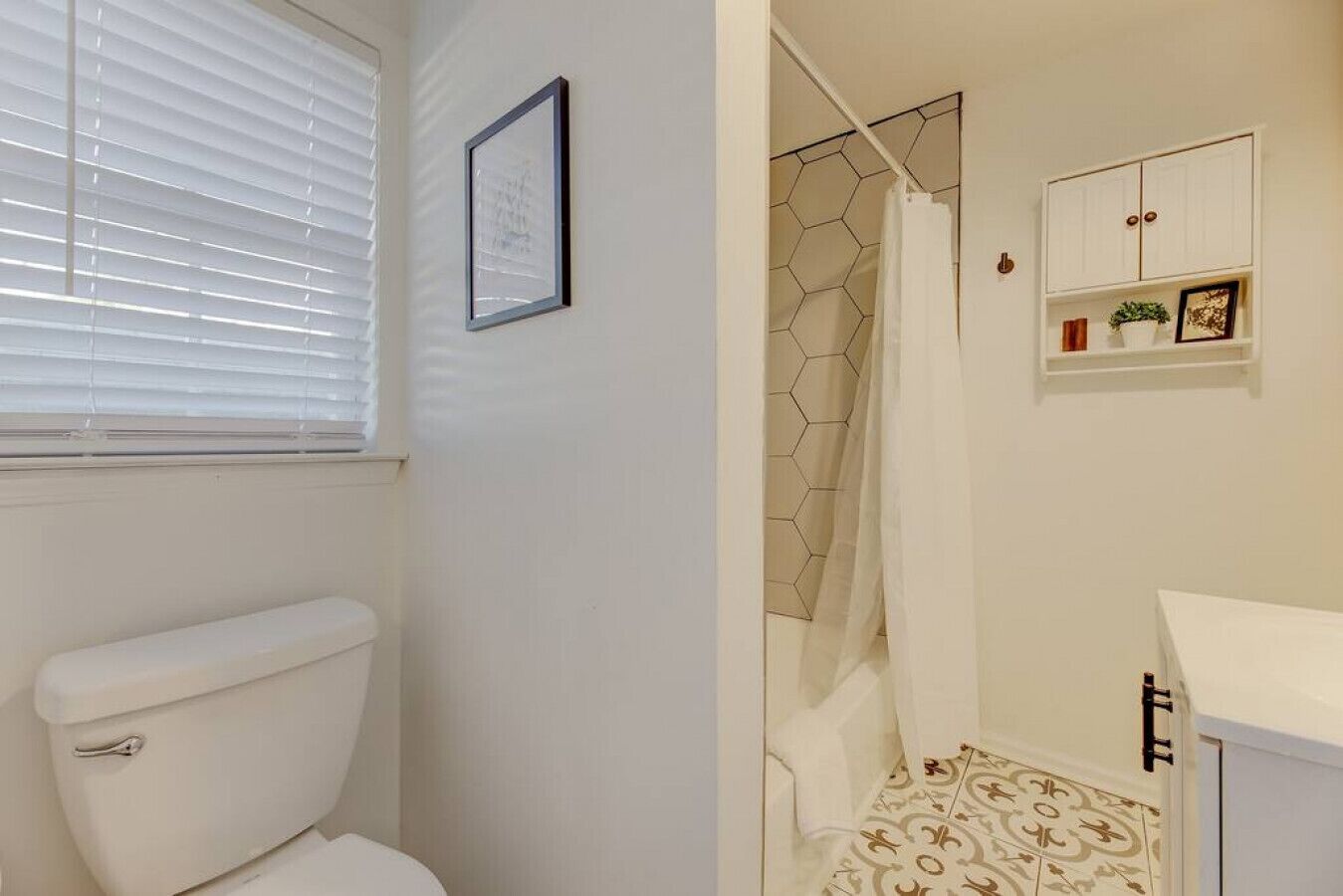 Une salle de bain comprenant des toilettes, une douche avec un mur recouvert de carreaux hexagonaux et un meuble blanc avec une étagère.