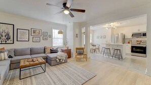 A modern living room with a grey sofa, a wooden coffee table, and a patterned rug. There is a ceiling fan, a dining area with a table and chairs, and a kitchen with white cabinets and appliances. - Modern 4 BR Apt - Mins to USC, ZOO & Ft Jackson (Columbia)