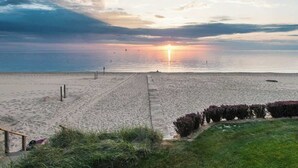 Una playa con un camino de madera que lleva al agua, un área con césped y arbustos, y un atardecer sobre el océano.