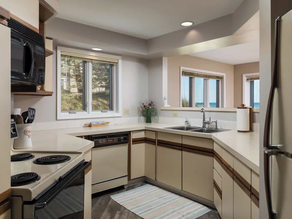 A modern kitchen with a dishwasher, stove, and refrigerator. There are windows providing natural light and a view of the outdoors.