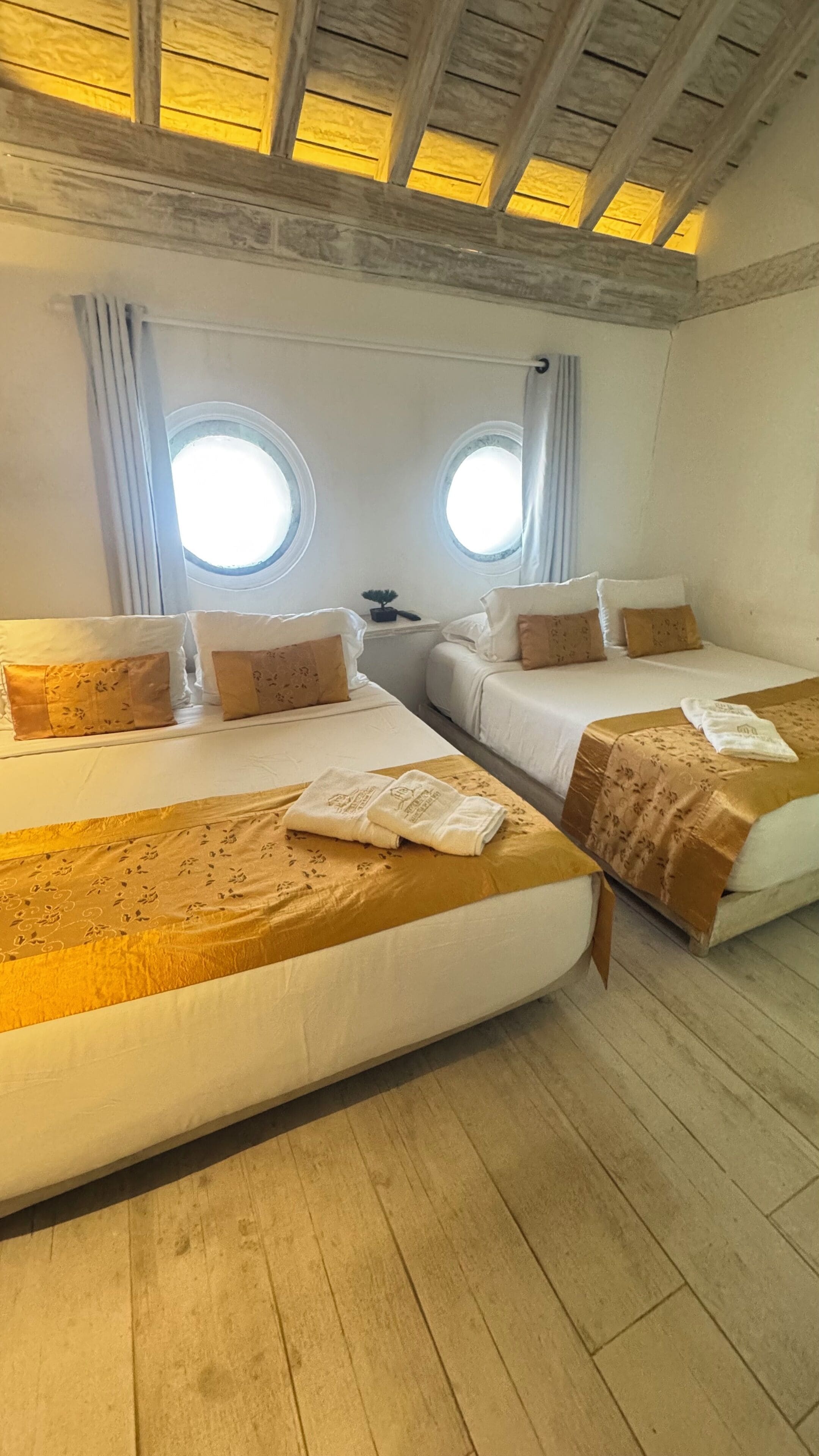 A hotel room with two beds, each with a headboard, a small table, and two windows.