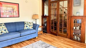 Een woonkamer met een blauwe bank, gekleurde kussens, een houten vloer, een glazen deur en een lijst met een afbeelding aan de muur.