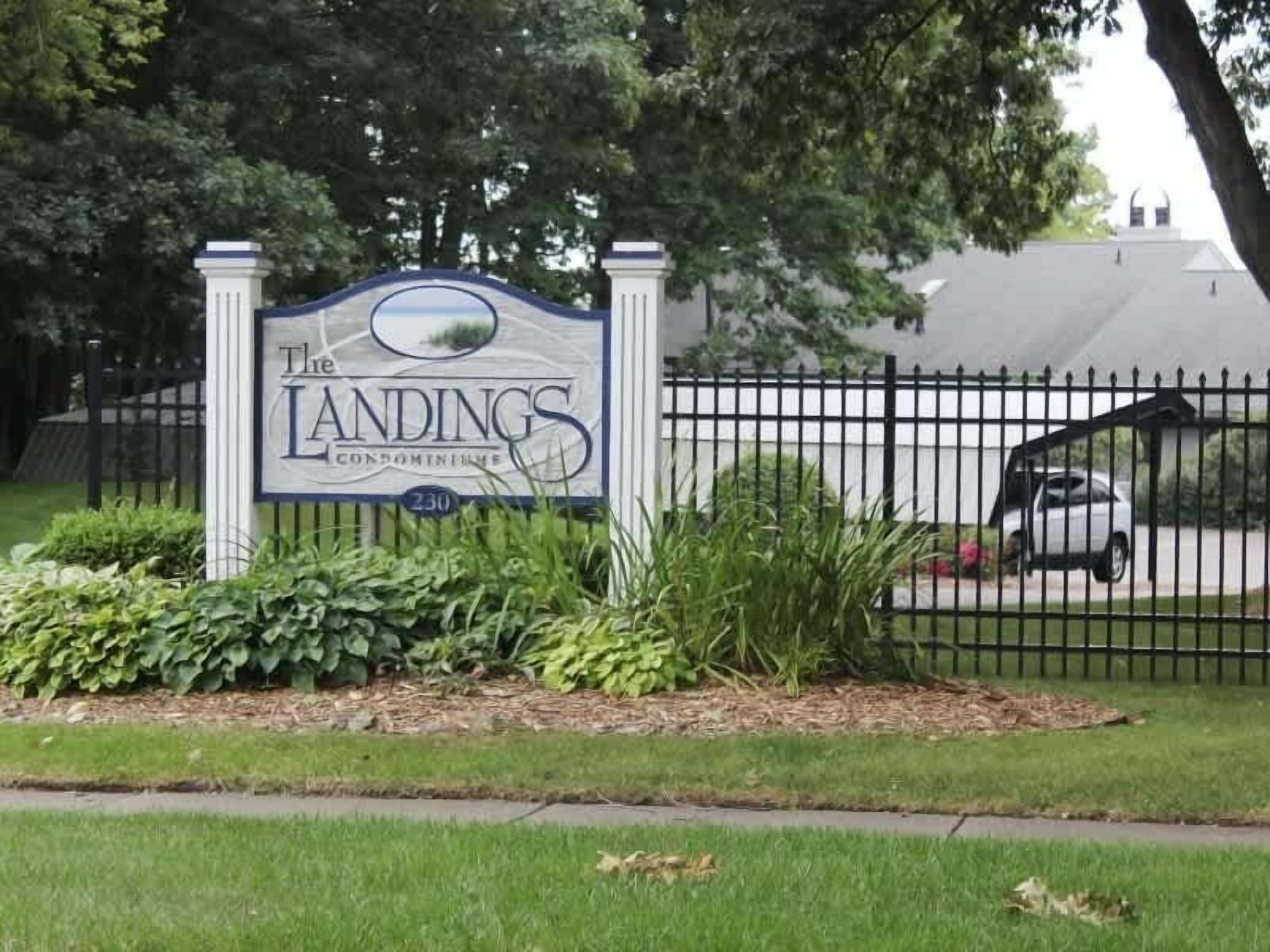 A sign for The Landings Condominium with a black fence and a car parked behind it.