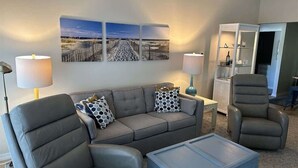 Een woonkamer met een hoekbank, een fauteuil en een salontafel. Er staat een dressoir met een lamp en een fotolijstje.